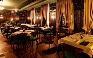 Alt tekst za ovu sliku: **Elegantno osvetljena unutrašnjost restorana Zlatni Bokal u Beogradu sa drvenim nameštajem, zavesama u toplim tonovima i stolovima pripremljenim za večeru**