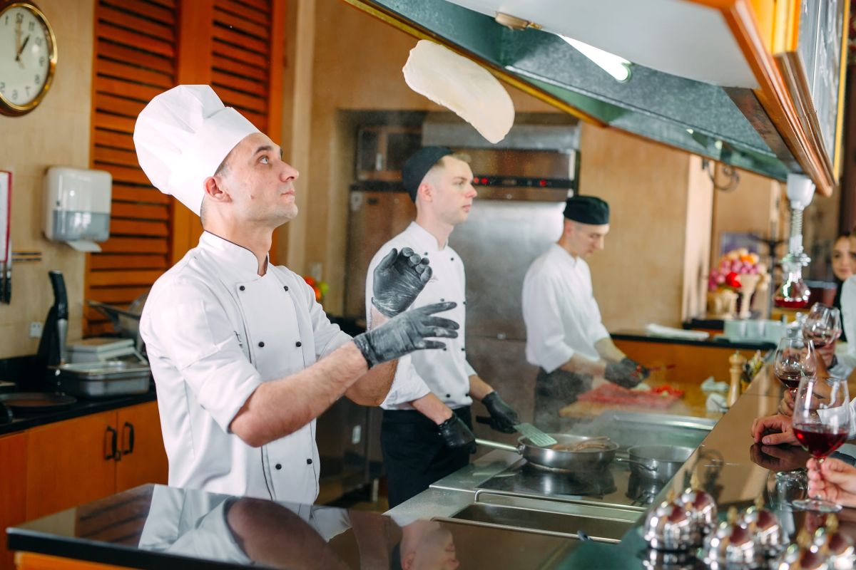 Kuvar u beloj uniformi baca testo u vazduh u otvorenoj restoranskoj kuhinji, dok ostali kuvari pripremaju jela.