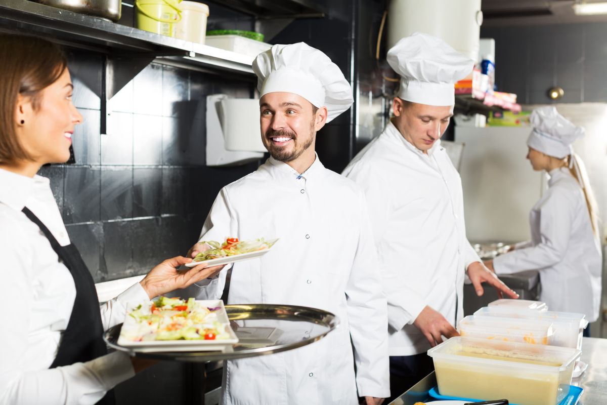 Kuvari u belim uniformama pripremaju jela u restoranskoj kuhinji, dok konobarica preuzima tanjire sa hranom za posluživanje