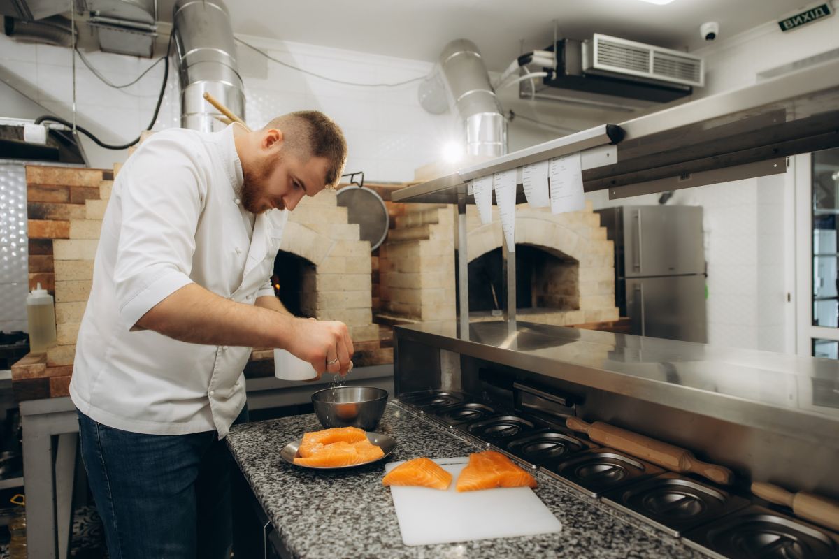 Kuvar u beloj uniformi priprema filete lososa na radnoj površini u profesionalnoj restoranskoj kuhinji